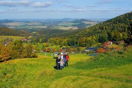 Wanderer auf einer grünen Wiese im Naturpark Zittauer Gebirge. Im Hintergrund grüne Berge und blauer Himmel.